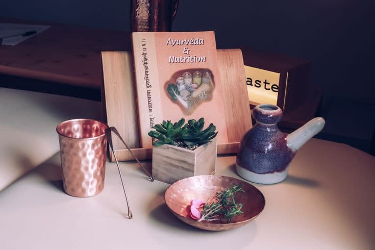 Deux gobelets en cuivre, un pot à huile, des plantes et un livre « Ayurvéda & Nutrition » posés sur une table